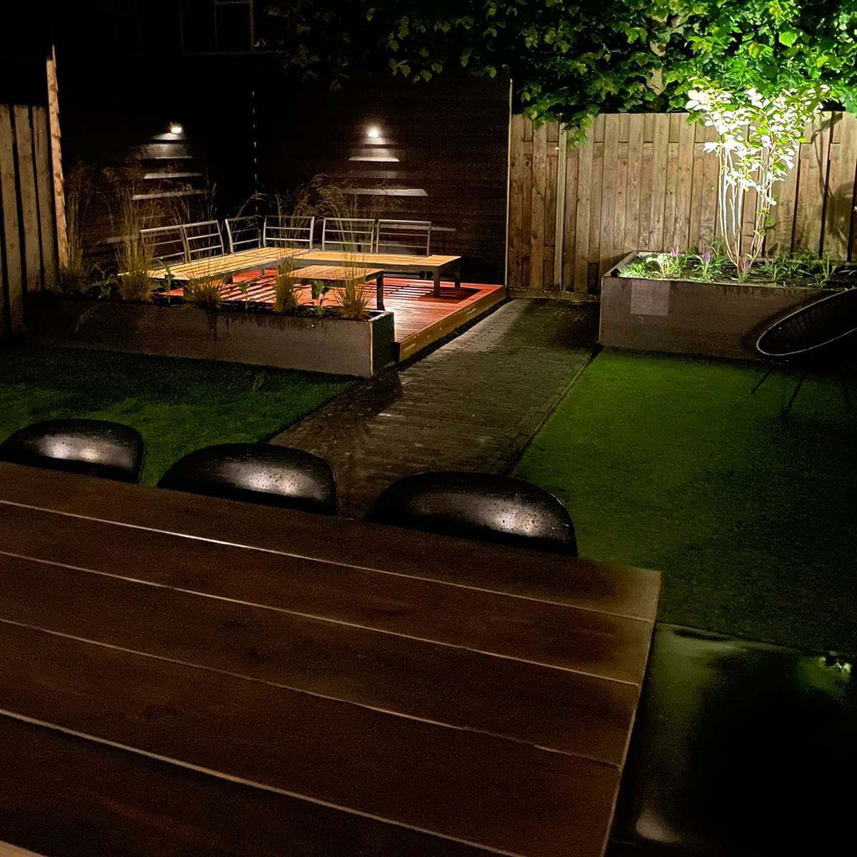 Tuin in de avond met verlichting, houten tafel, kunstgras en plantenbakken