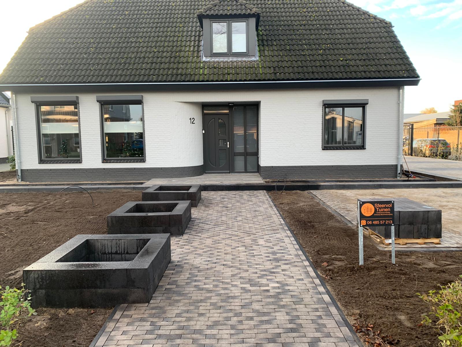 Nieuw aangelegde voortuin met grijze bestrating en zwarte plantenbakken, met het bord van Sfeervol Tuinen