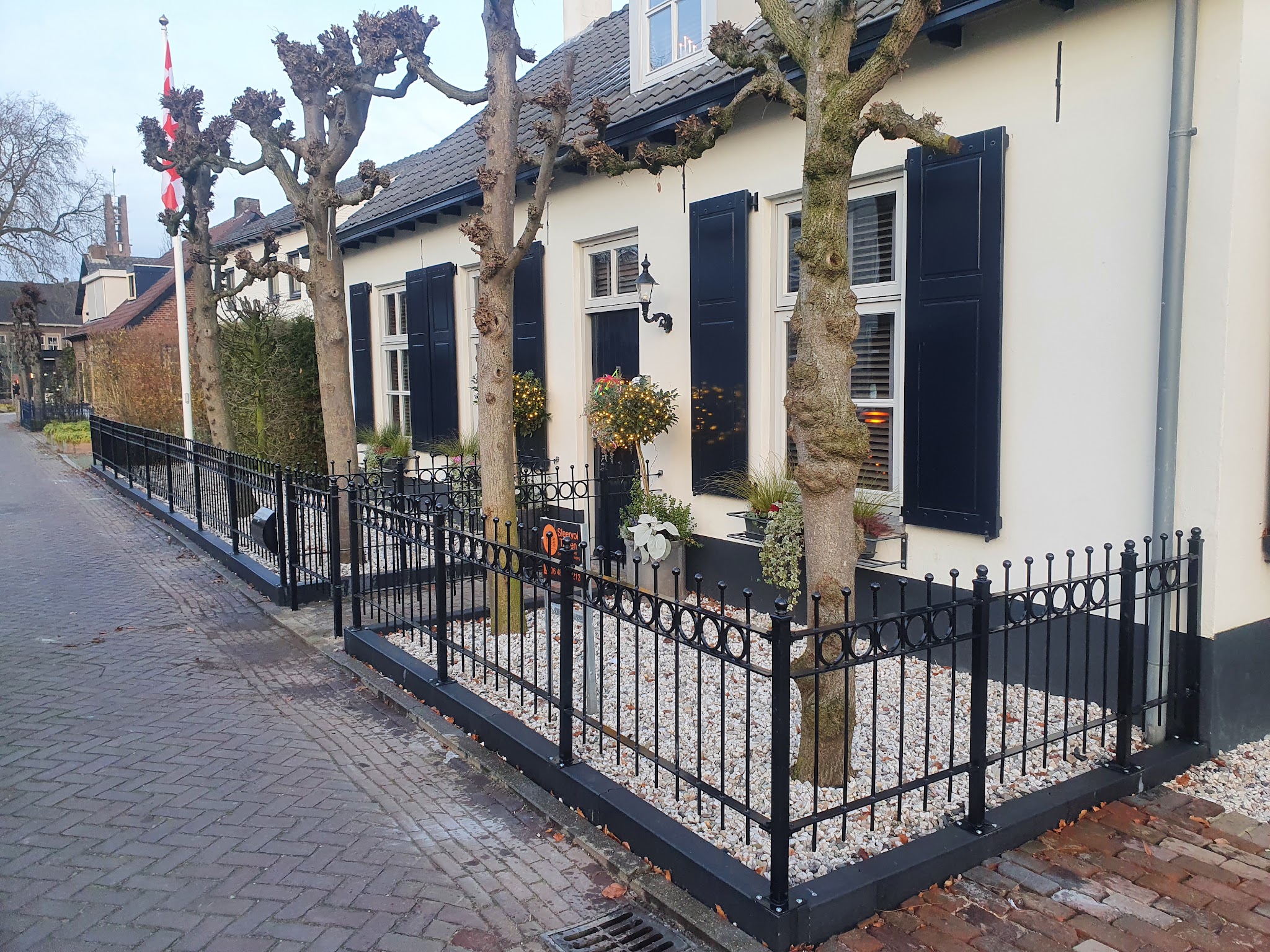 Voortuin met zwart smeedijzeren hekwerk, leibomen en wit grind voor een wit huis